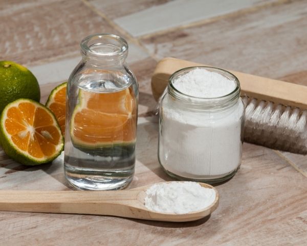 Ingredientes comuns usados em receitas de clareamento caseiro, incluindo bicarbonato de sódio em pote e colher, limão cortado e frasco com líquido transparente, dispostos sobre uma superfície de madeira.