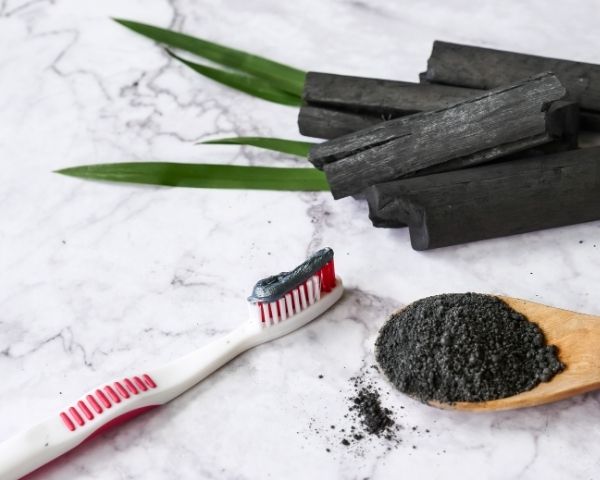 Carvão ativado em pó sobre colher de madeira e escova de dentes com o produto aplicado, representando um método popular de clareamento caseiro, embora ineficaz e prejudicial ao esmalte dental.